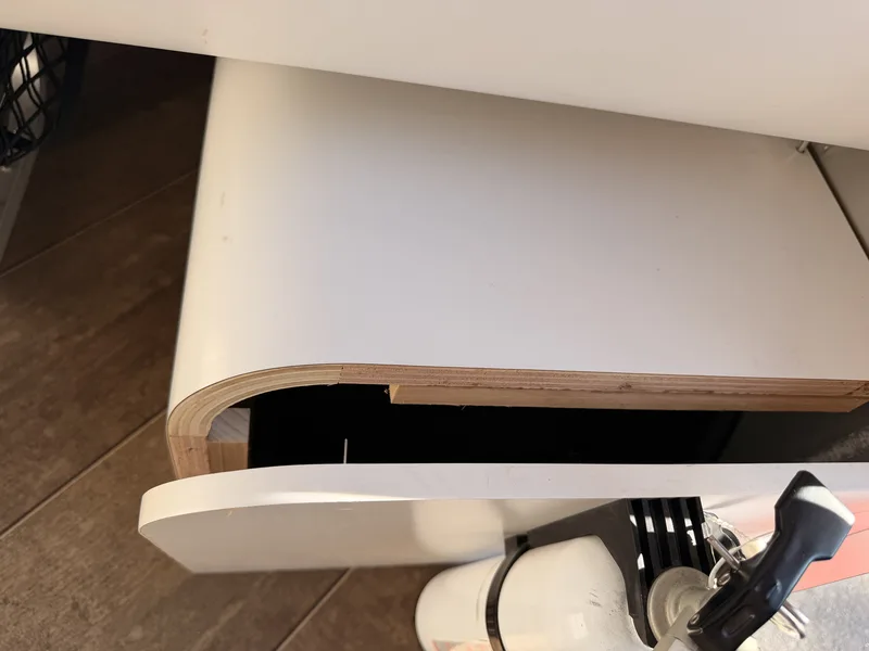 Close-up of a curling white laminate panel peeling back from its plywood core inside the Airstream near the propane tank