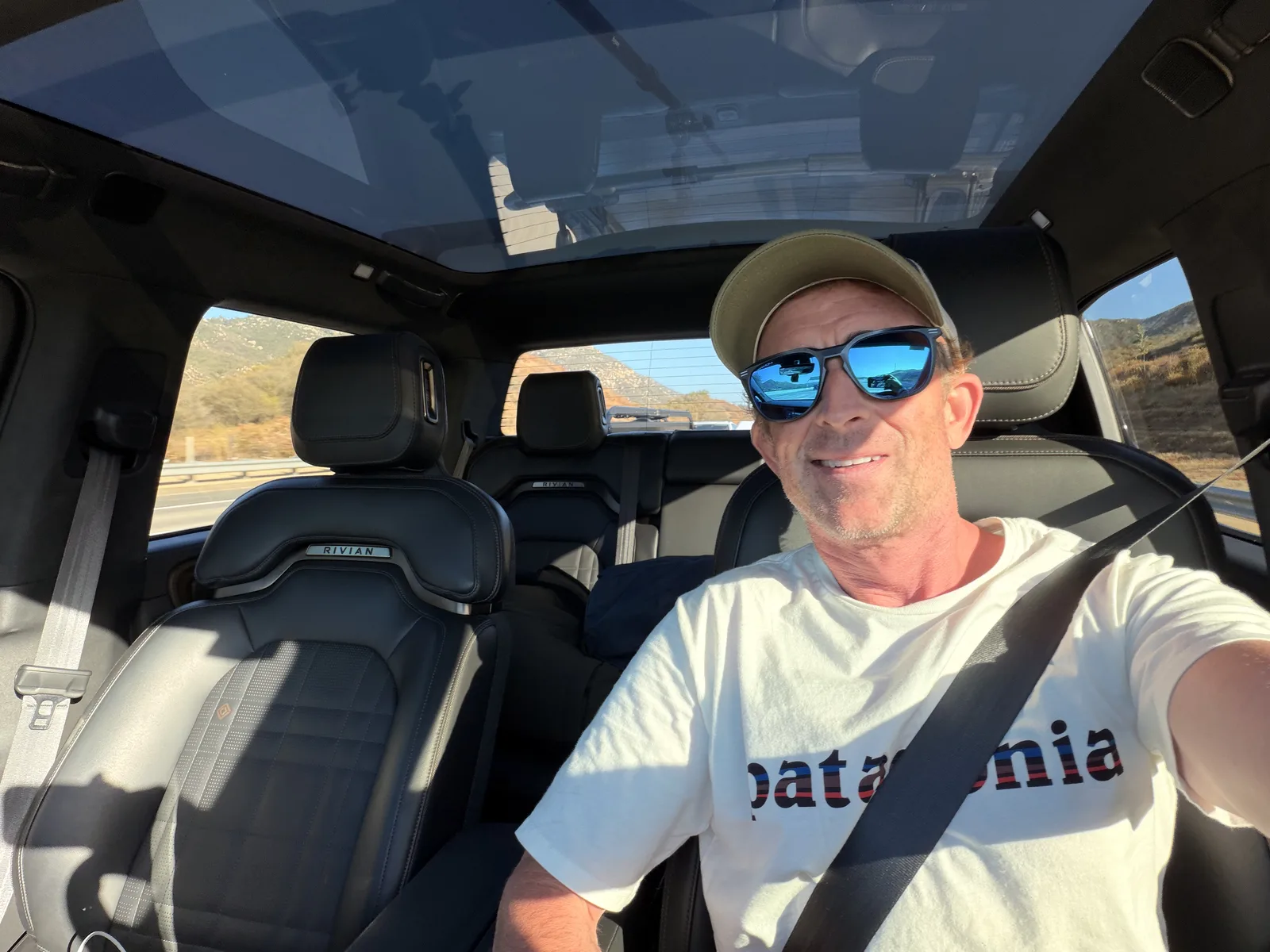 A grinning, sunglassed driver in a Patagonia tee in the front seat of the white Rivian R1T at the start of the Utah Ducati Run, late-afternoon light through the windshield.