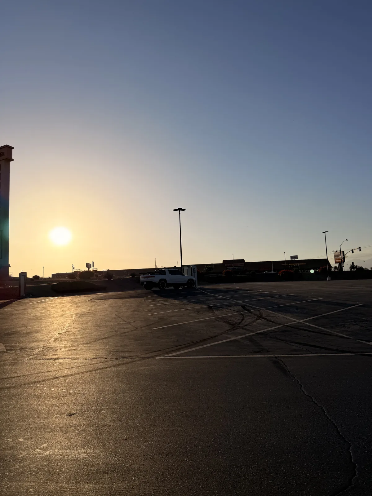 The white Rivian R1T parked across a near-empty lot as the sun drops to the horizon over the asphalt at the first charge stop of the Utah Ducati Run.