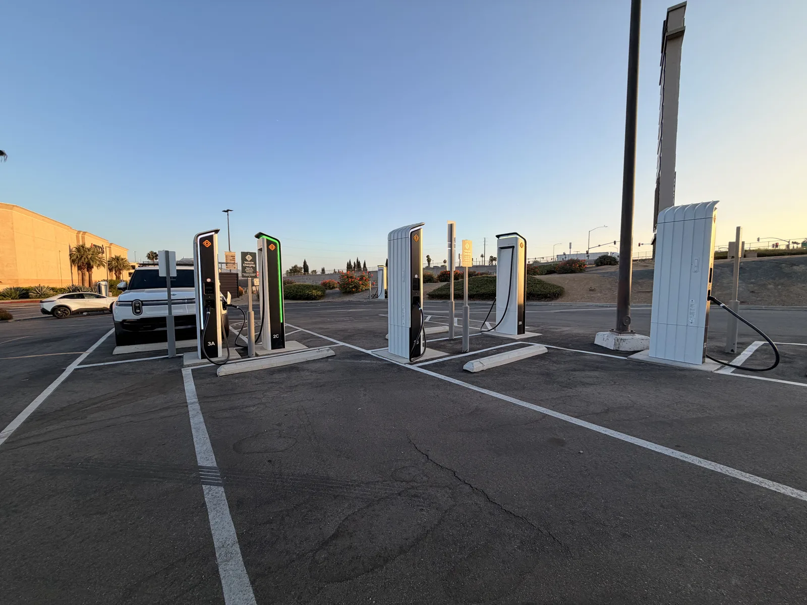 The San Bernardino Rivian Adventure Network charging plaza at golden hour, several white RAN pedestals with the Rivian R1T charging at the far stall, palms and the mall behind.