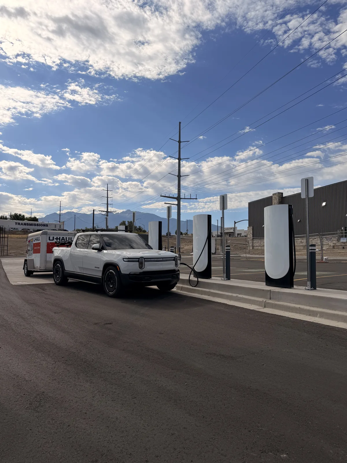 The white Rivian R1T charging at a Rivian Adventure Network pedestal in Sandy, Utah with a U-Haul rental depot behind, topping off before hitching the trailer.