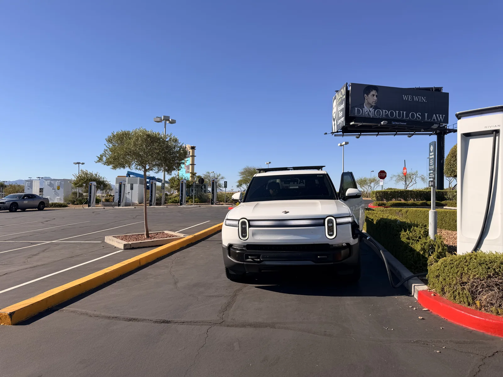 The white Rivian R1T charging at a Rivian Adventure Network pedestal in Las Vegas under a clear blue morning sky, palms and a billboard behind, on Day 4 of the Utah Ducati Run.