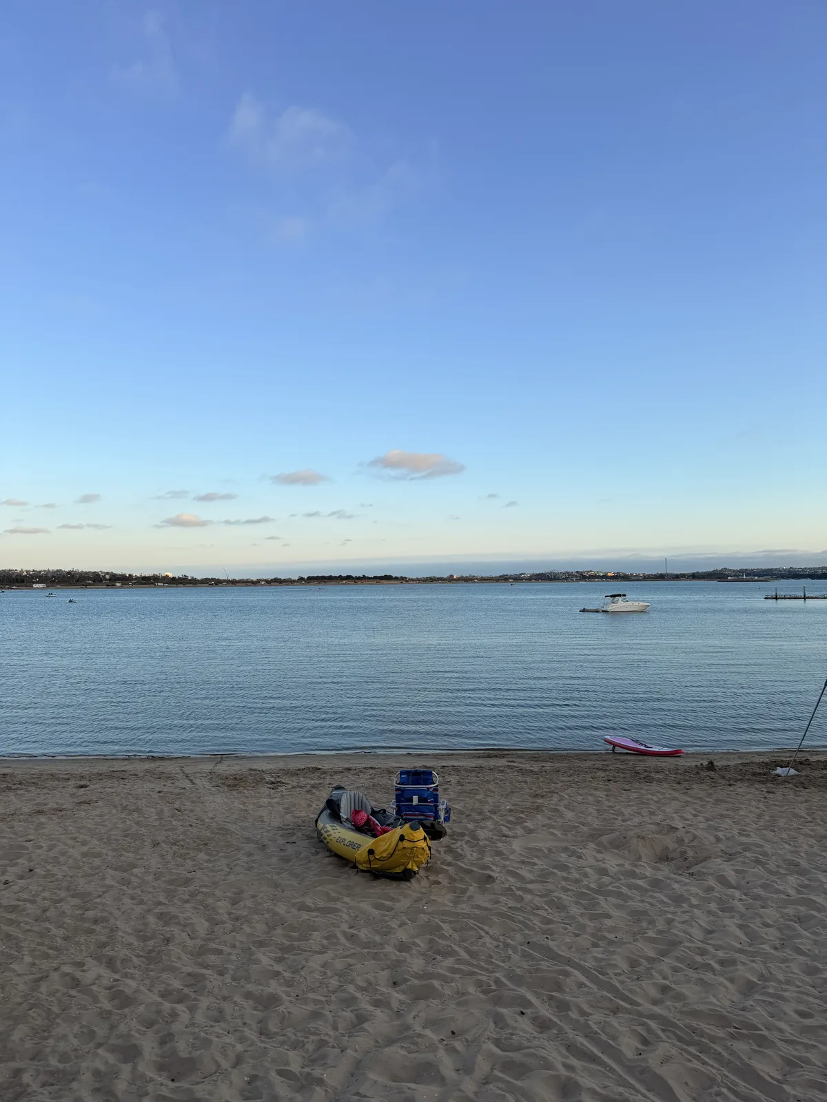 Mission Bay at golden hour from the beach at Campland on the Bay, a yellow kayak and a cooler on the sand and a boat at anchor on the flat water