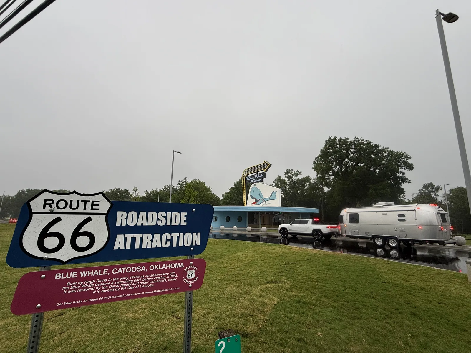 A "Route 66 Roadside Attraction, Blue Whale, Catoosa, Oklahoma" interpretive sign in the foreground, with the blue whale sculpture and the white Rivian R1T towing the silver Airstream lined up along the pond behind it on a wet overcast morning