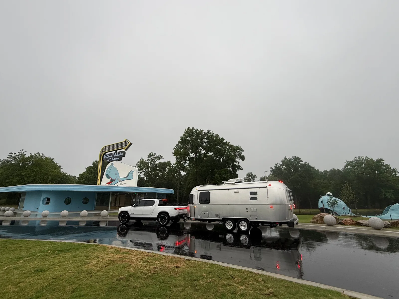 The light-blue concrete sperm-whale sculpture resting in its green spring-fed pond, the white Rivian R1T and silver Airstream parked on the access road alongside, reflections in the rain-wet pavement, leafy trees and overcast sky