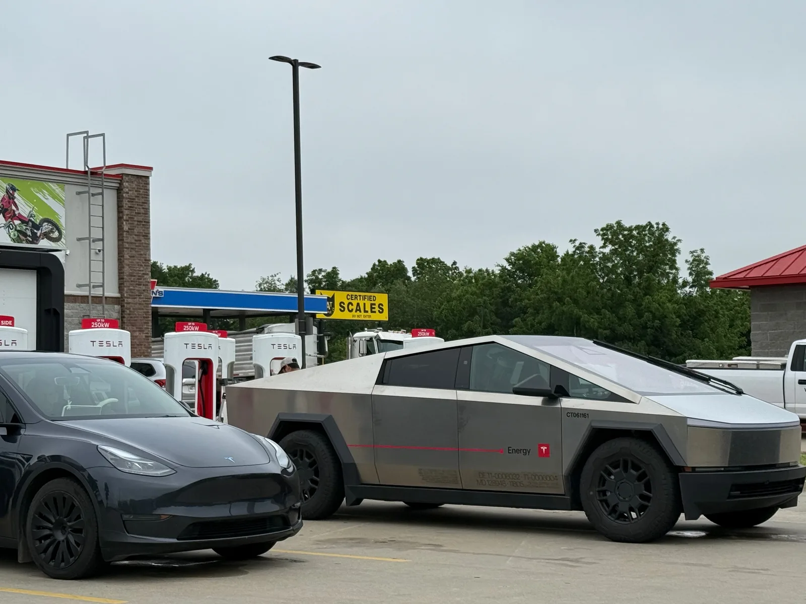 A stainless-steel Tesla Cybertruck and a dark Tesla Model Y charging at the Lowell AR Supercharger, the red-and-white stalls and a Love's-style travel stop behind under an overcast sky