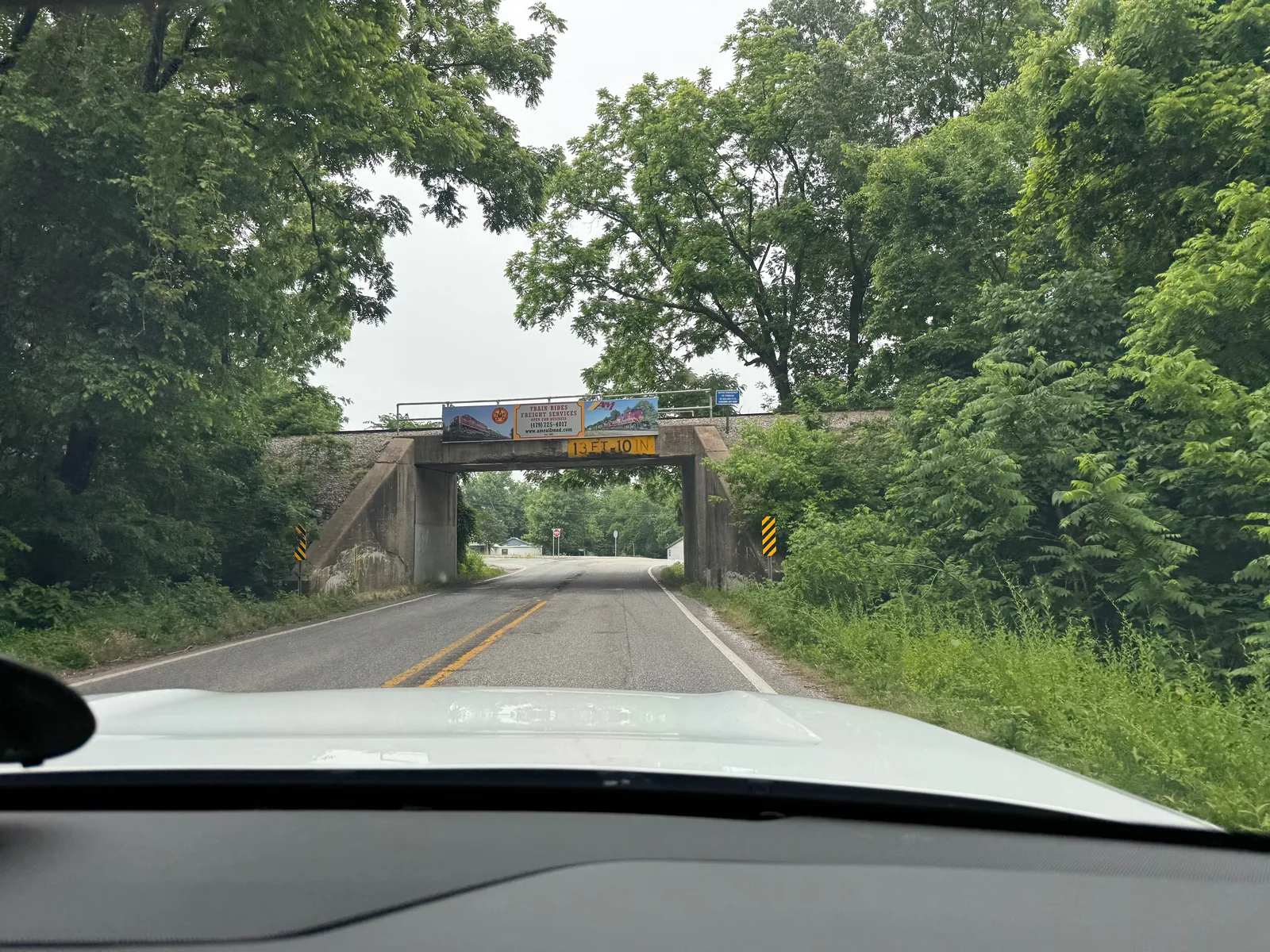 The view through the Rivian windshield on a two-lane road approaching a stone railroad underpass in the green Ozark woods, heading north from Arkansas toward Missouri