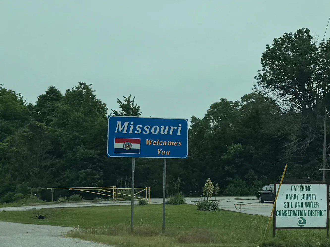 A blue "Missouri Welcomes You" state-line sign with the Missouri state seal beside a wooded two-lane highway, a soil-and-water conservation district sign next to it, overcast Ozark sky