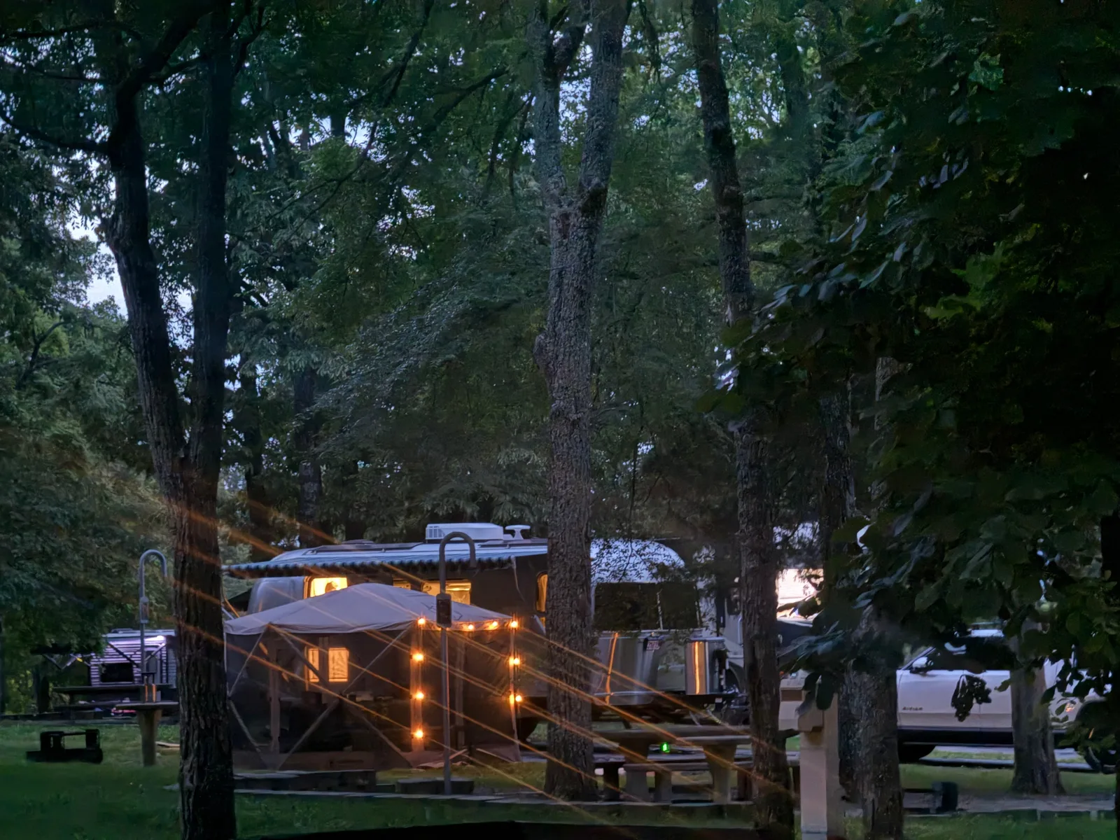 The silver Airstream International glowing warm from its interior lights at dusk among the trees at the Indian Point campsite, deep-blue evening sky behind