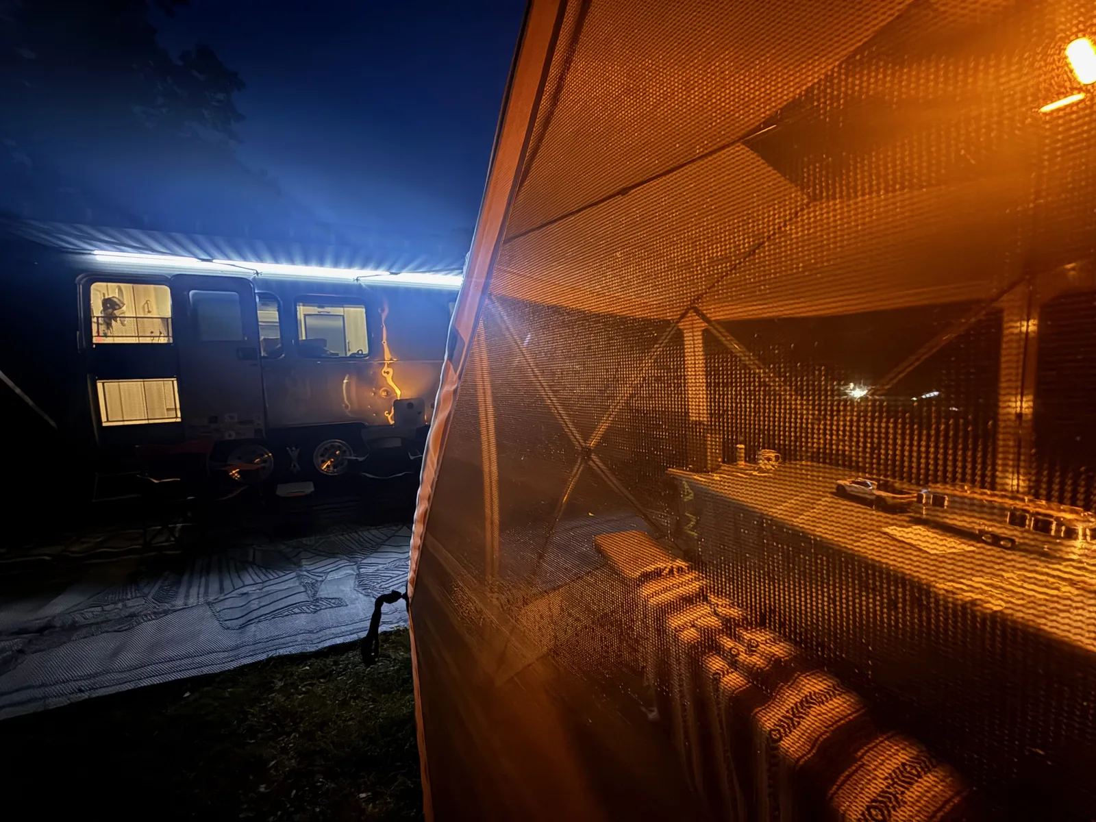 Inside the Gazelle screen gazebo at night, string lights glowing warm overhead, the lit Airstream visible through the mesh wall at the Indian Point campsite