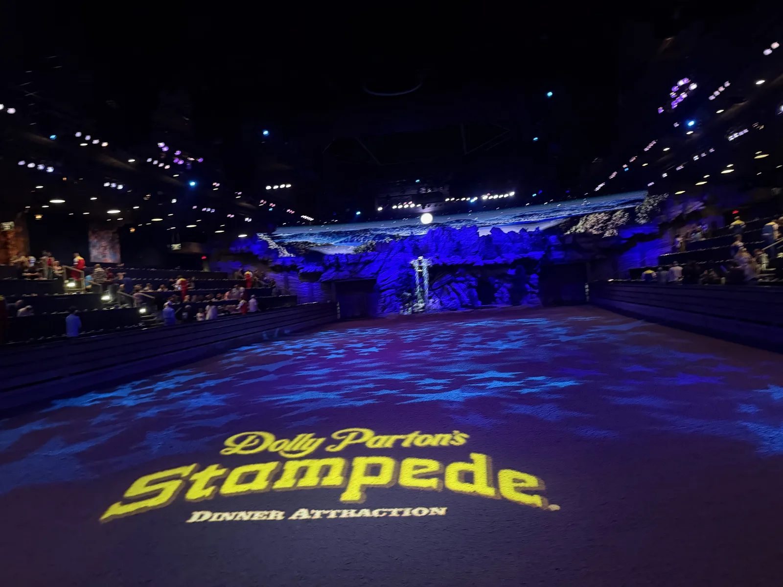 The dirt arena floor at Dolly Parton's Stampede lit deep blue with "Dolly Parton's Stampede, Dinner Attraction" projected onto the ground before the show