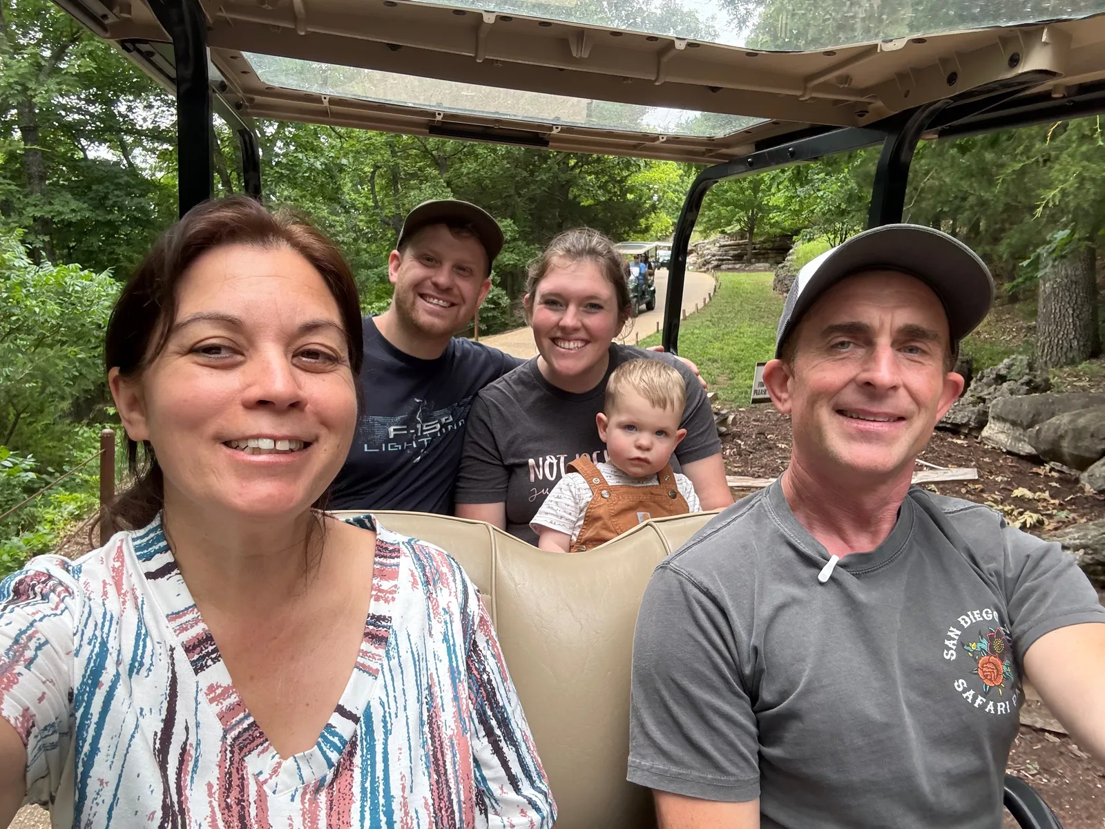 Diana and Eric smiling in a selfie from the front of the golf cart on the Lost Canyon Cave trail, green Ozark woods behind