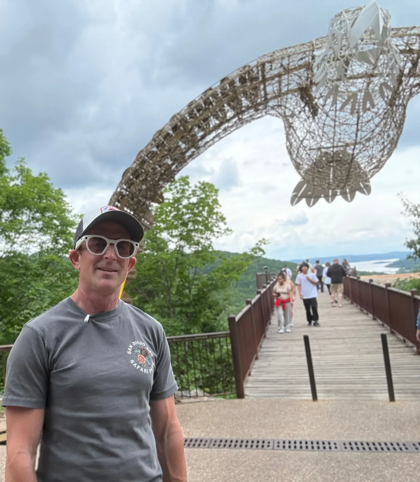 Eric on the Top of the Rock boardwalk overlook beneath a giant open-frame steel fish sculpture, Table Rock Lake and the Ozark hills falling away behind