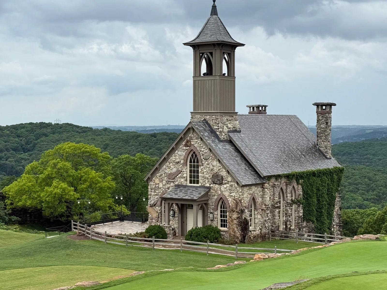A small stone chapel with a steeple on a green bluff at Top of the Rock, the rolling Ozark hills behind under a cloudy sky