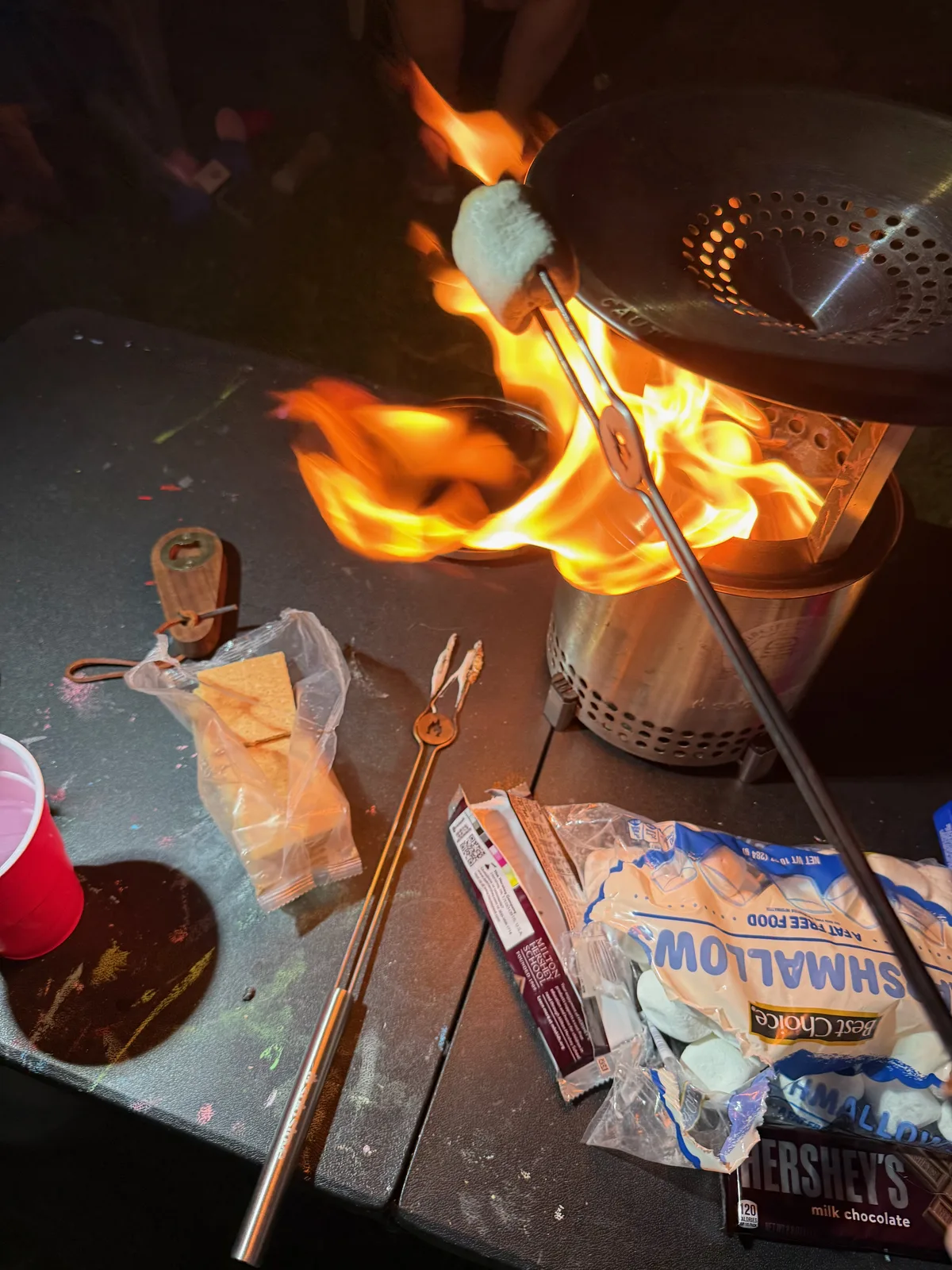 A marshmallow on a long roasting fork held over the flames of a Solo Stove Mesa XL fire pit at night, with a bag of marshmallows, a Hershey's milk chocolate bar, and a red Solo cup on the table beside it, the makings of s'mores at the farm