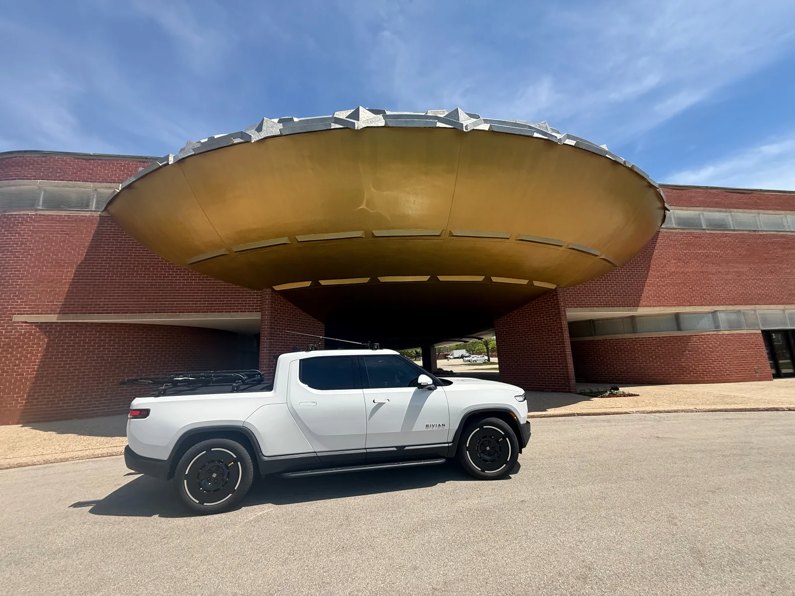The white Rivian R1T parked beneath the gold saucer-shaped roof of the Golden Rondelle Theater on the SC Johnson campus in Racine, Wisconsin, red brick walls around it