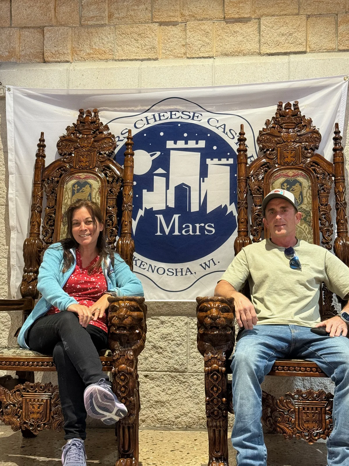 Two travelers seated in oversized carved wooden thrones in front of a Mars Cheese Castle, Kenosha Wisconsin banner