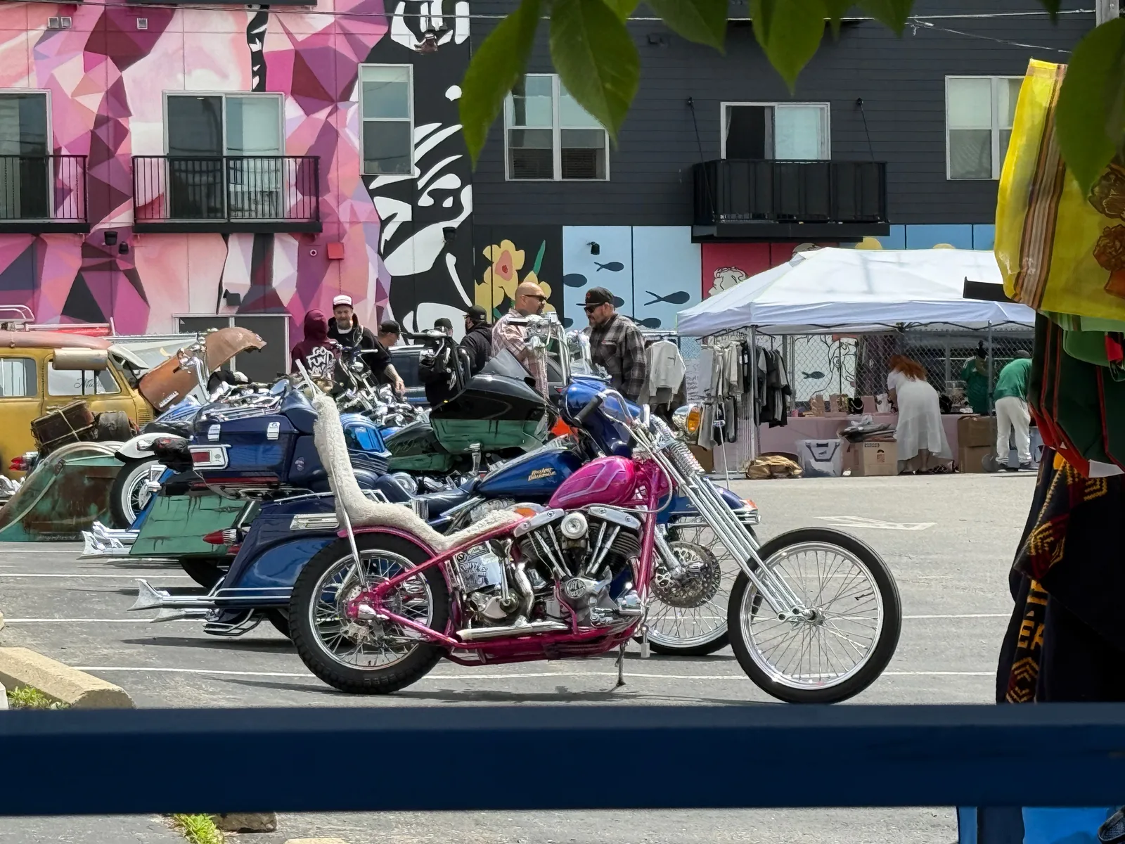 A row of custom motorcycles in a Milwaukee lot against a wall of bright murals, a pink hardtail chopper in front of baggers and a vintage truck, vendor tents along the fence