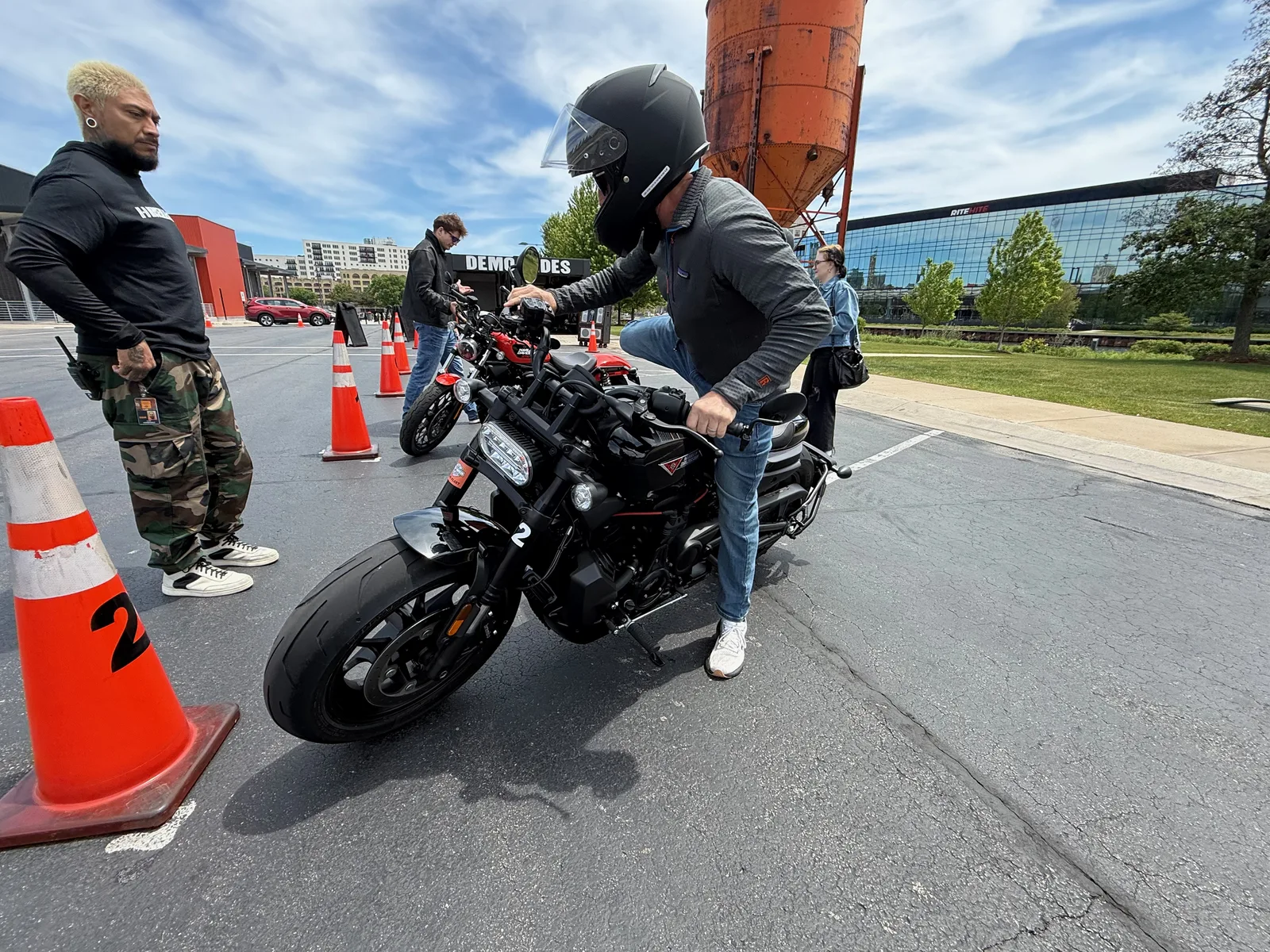 A rider in a black helmet mounting a red Harley-Davidson Sportster at the cone-lined demo-ride course on the Harley-Davidson Museum campus, an attendant standing by
