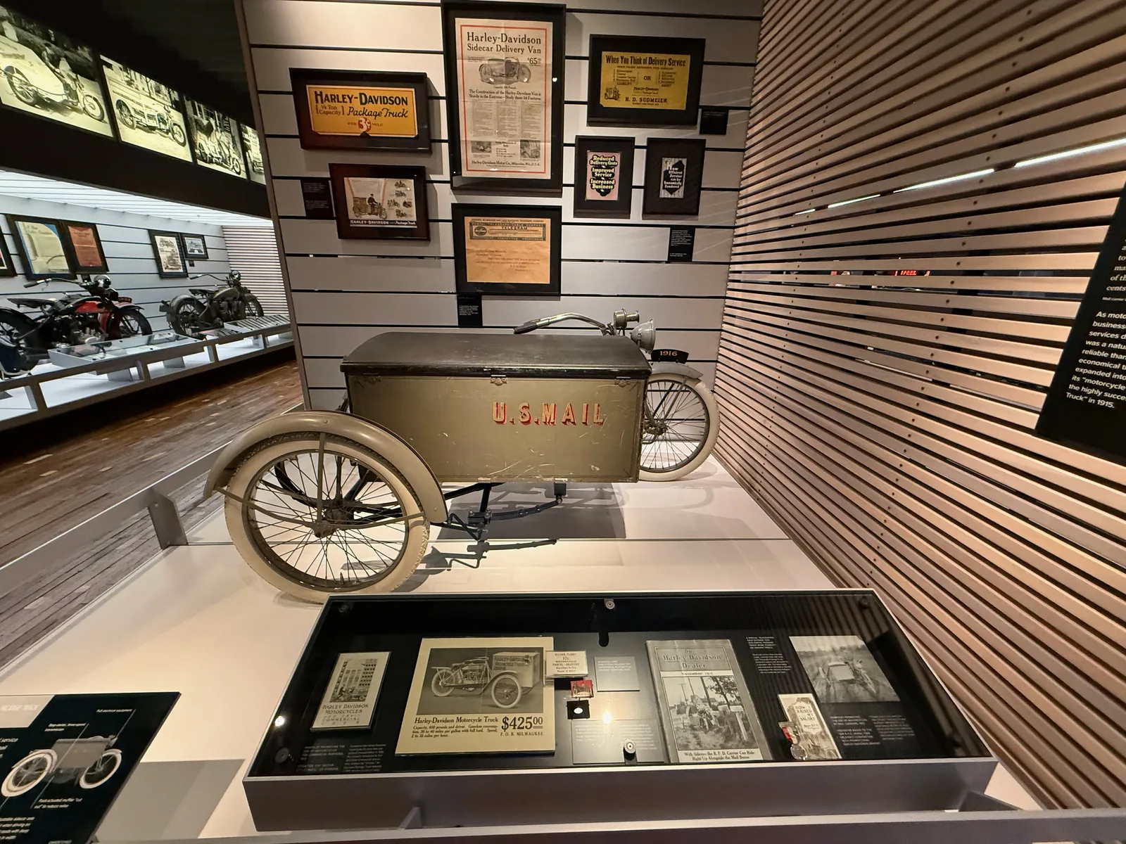 A vintage U.S. Mail Harley-Davidson sidecar delivery truck with a green cargo box, displayed below a wall of early Harley package-truck advertisements