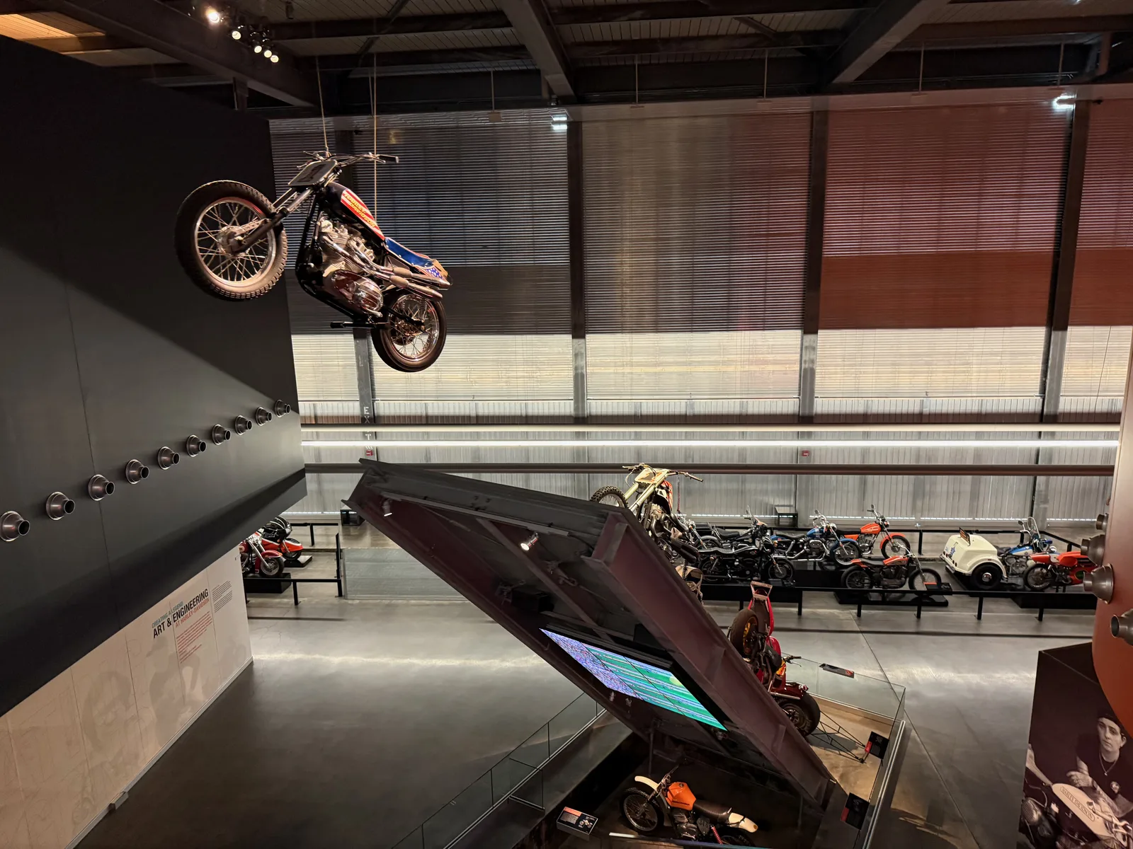 The soaring Art and Engineering atrium at the Harley-Davidson Museum with a motorcycle mounted climbing the wall above rows of bikes on the floor below
