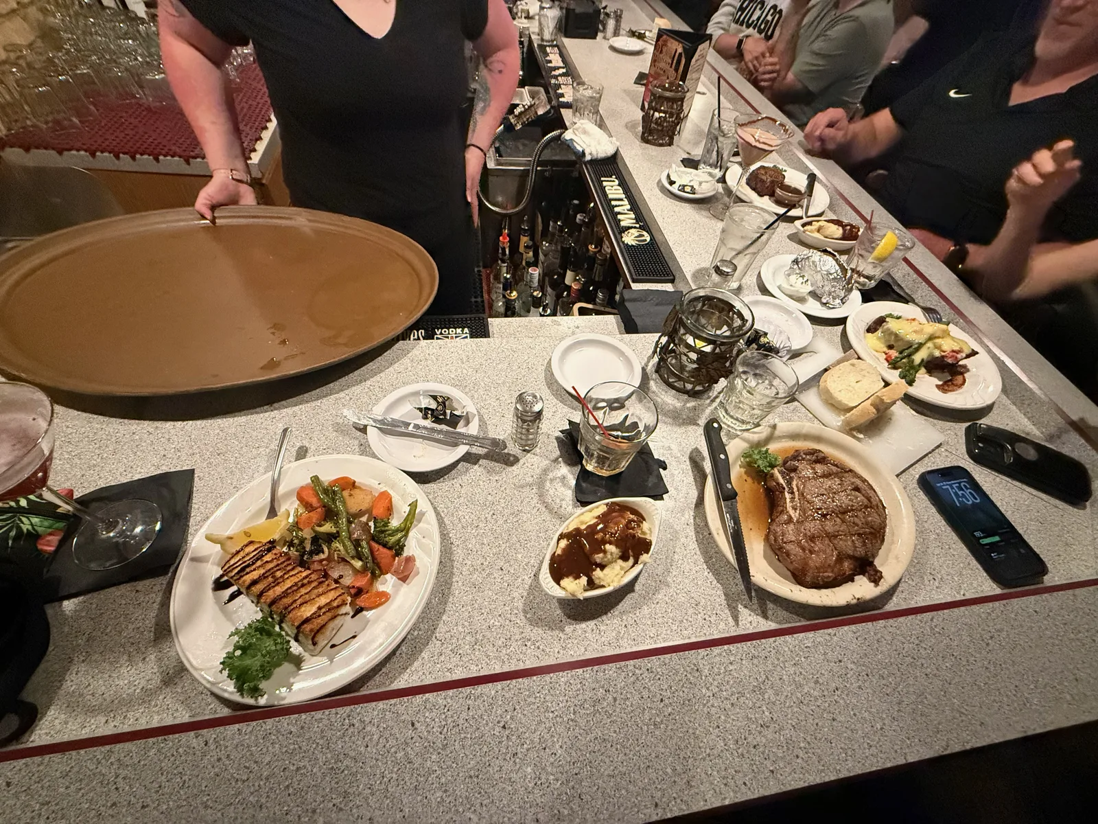 A spread of supper-club plates on the bar at Colony House, Eric's ribeye steak, Diana's halibut, a grilled vegetable plate and mashed potatoes, a server carrying a tray