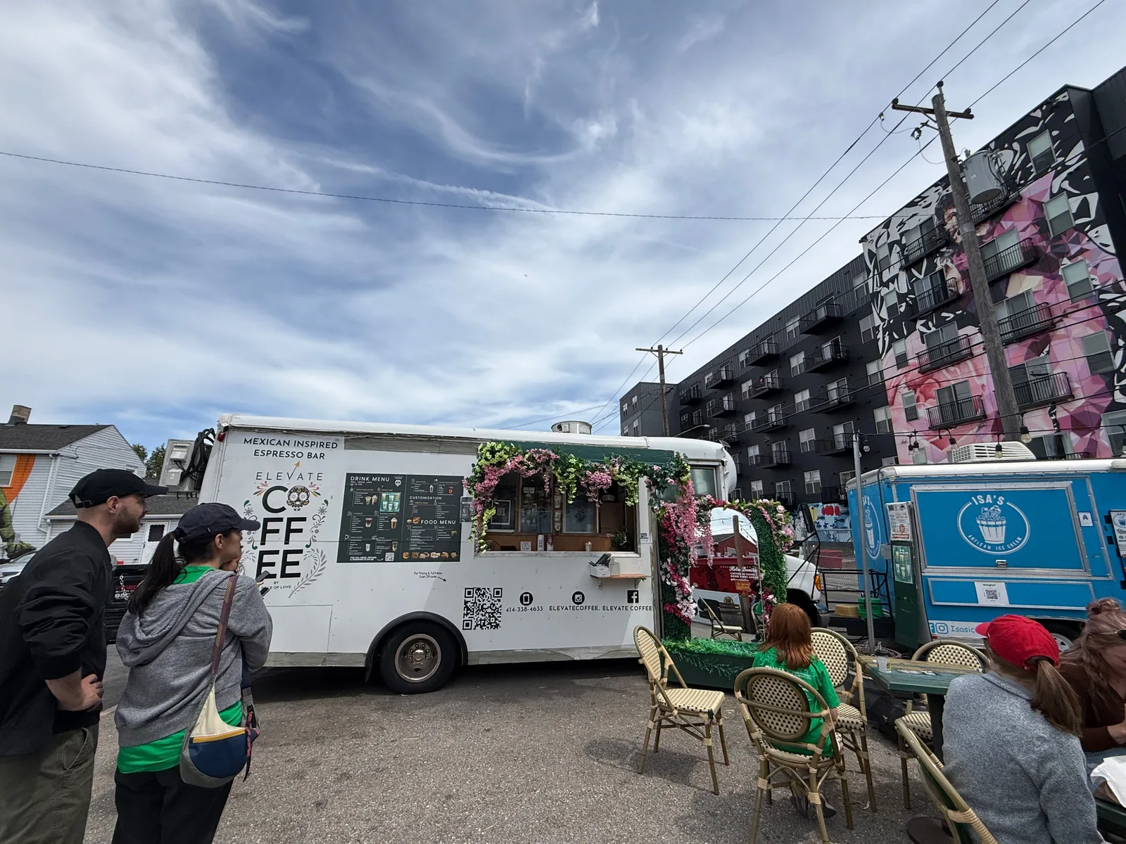 Food trucks at an open-air park near Milwaukee's Historic Third Ward, Elevate Coffee's Mexican-inspired espresso bar truck and a blue Isa's juice truck against a wall of murals