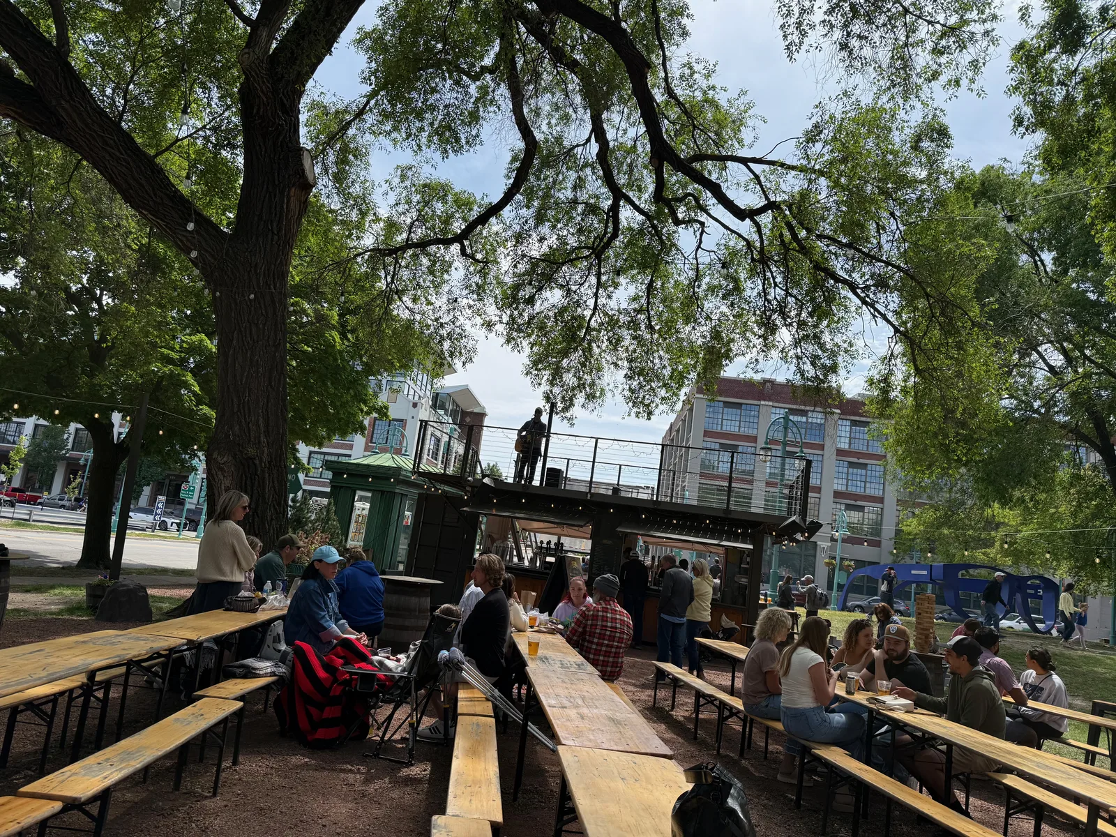 A shaded Milwaukee beer garden under big oak trees with long communal picnic tables and people gathered, brick buildings beyond