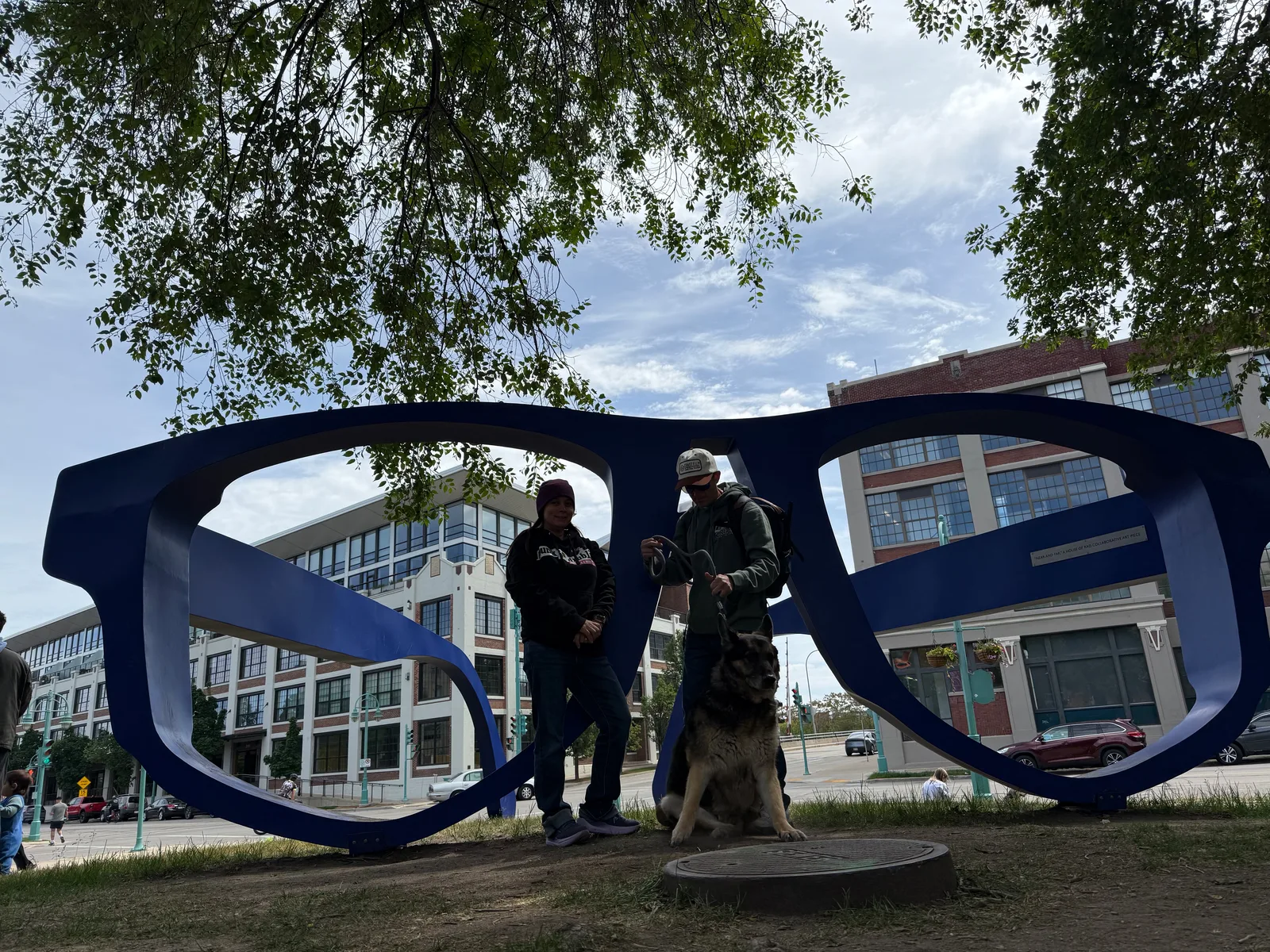 A person and Mission the German shepherd posing inside an oversized blue eyeglasses sculpture in Catalano Square in Milwaukee's Historic Third Ward, brick warehouses behind