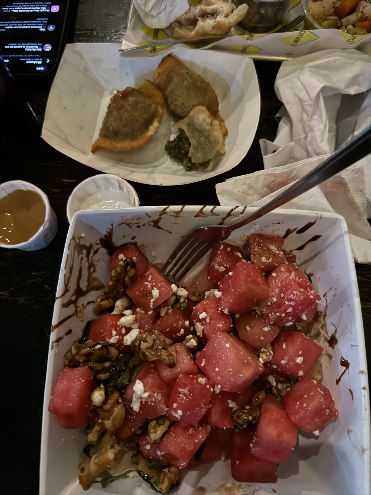 A watermelon, feta, and candied-walnut salad with balsamic in a white bowl, fried empanadas with pesto on a tray behind it