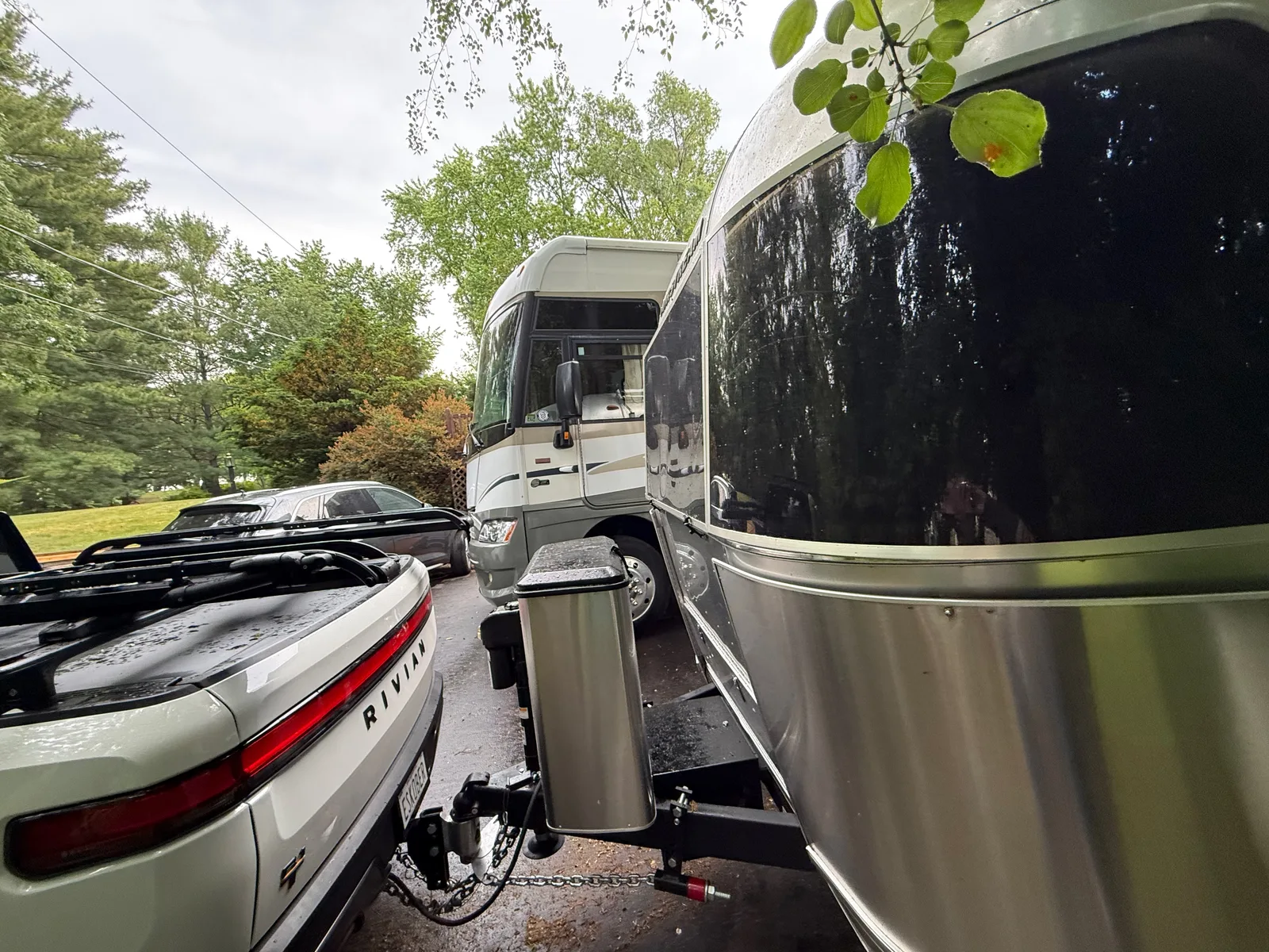 The white Rivian R1T hitched to the polished Airstream, parked beside a motorhome under green trees on a damp morning near Fox Lake