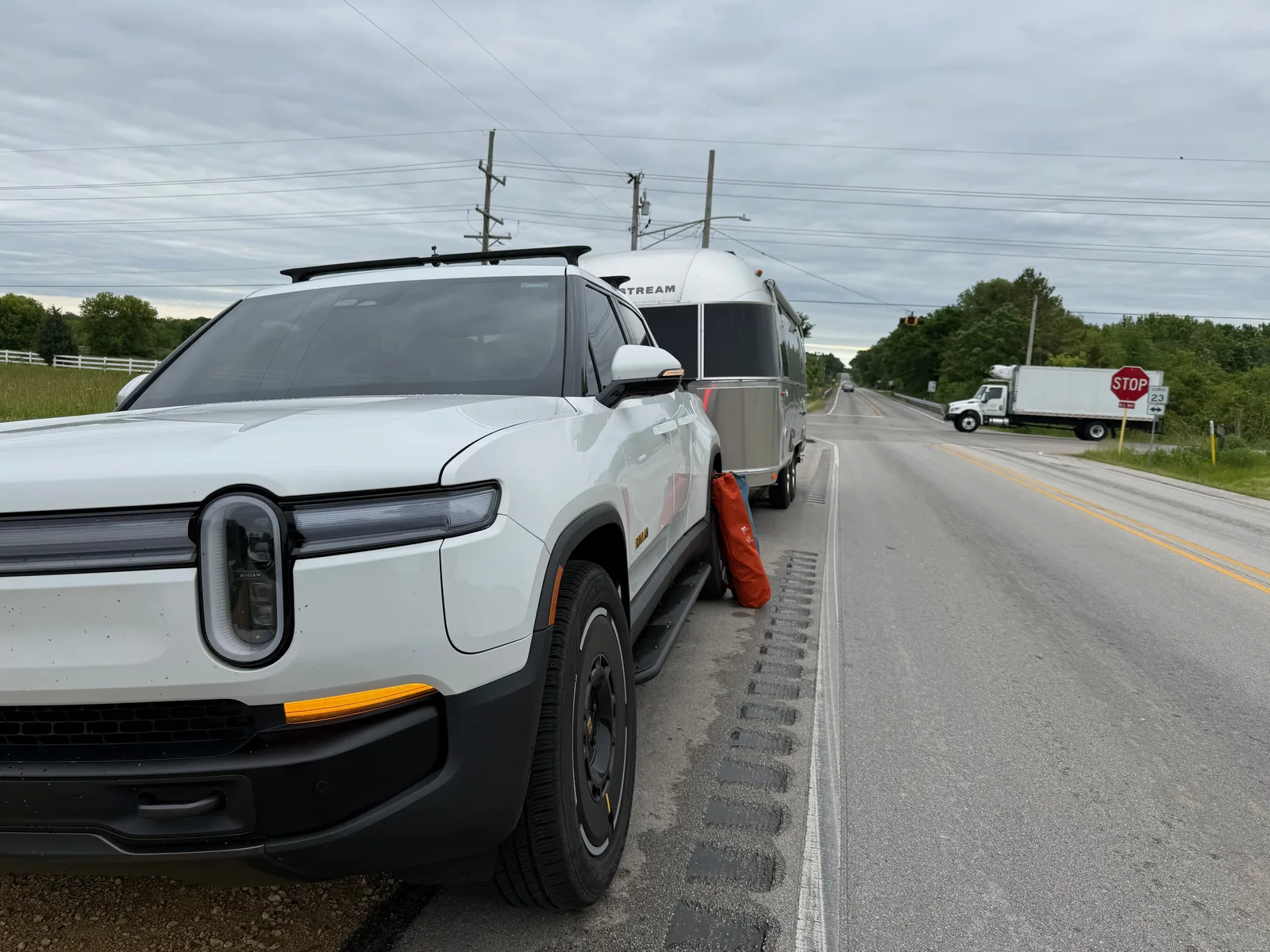 The white Rivian R1T and hitched Airstream pulled onto a rural highway shoulder on a cloudy morning, a red gas station across the two-lane road