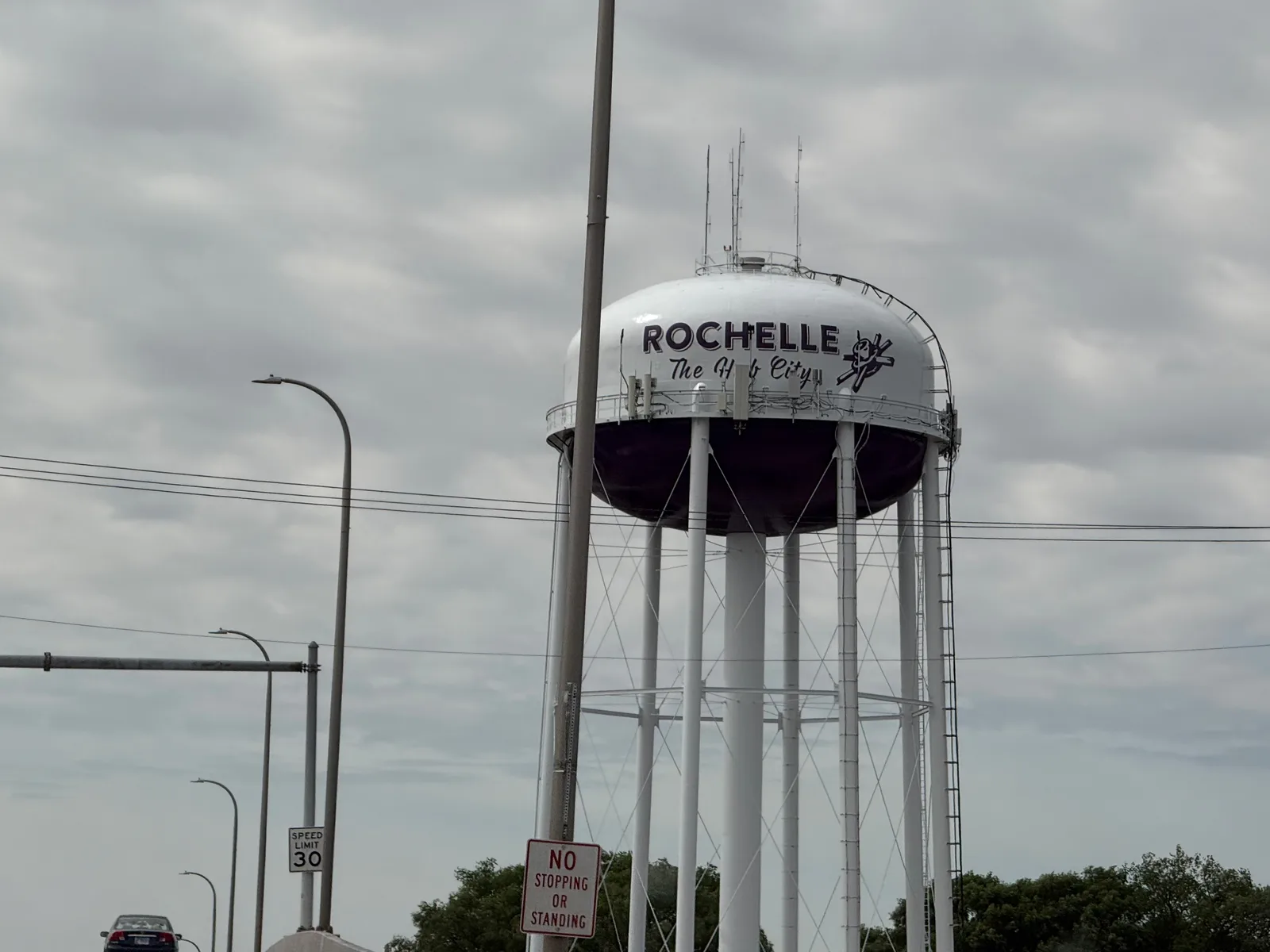 The Rochelle, Illinois water tower painted "Rochelle, The Hub City" with a railroad-crossing logo, seen against an overcast sky from the highway below
