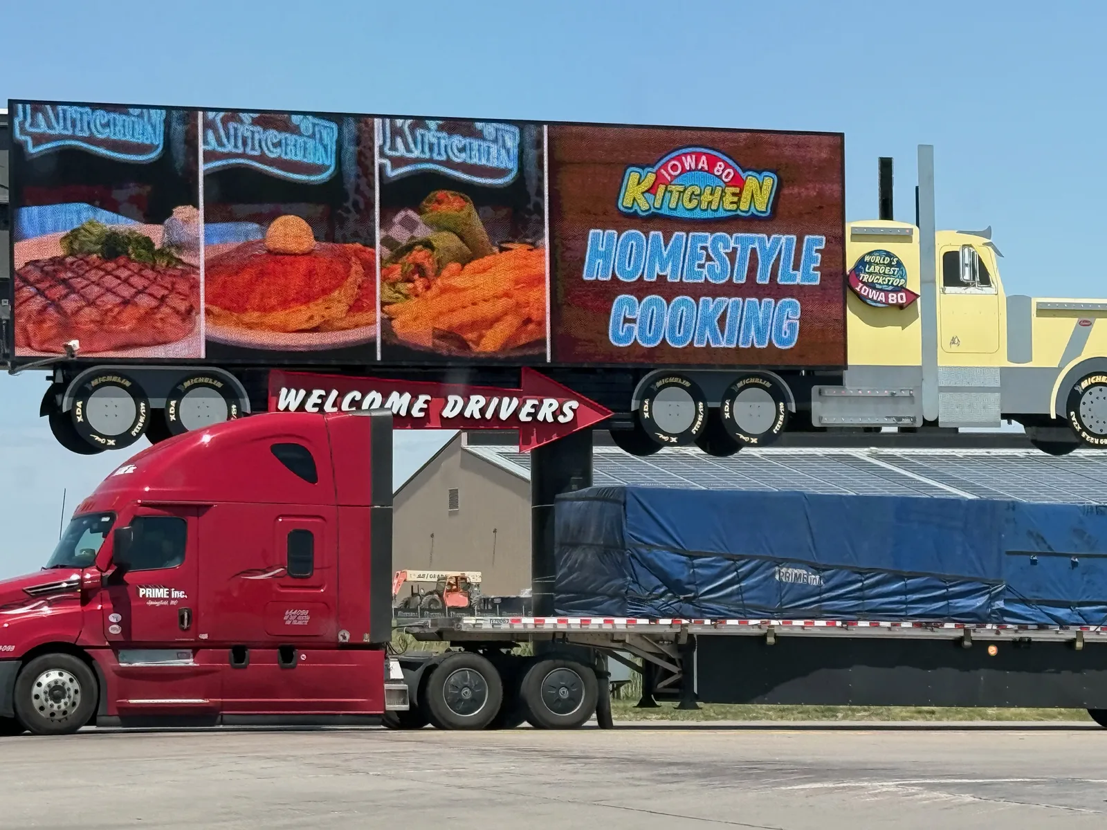 The Iowa 80 Truckstop entrance arch reading "Welcome Drivers" and "Iowa 80 Kitchen Homestyle Cooking" with a red Prime semi pulling a flatbed underneath