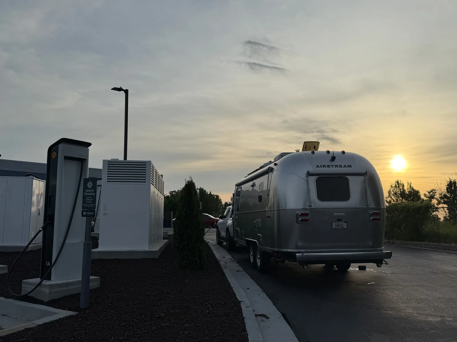 Polished Airstream hitched to the white Rivian R1T at a Rivian Adventure Network pedestal as the sun rises over Independence, Missouri