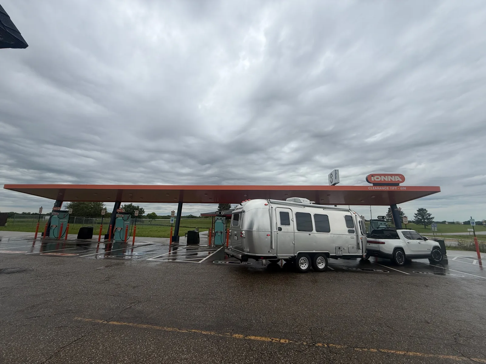 The Airstream and Rivian parked under the long orange IONNA canopy on a wet lot under heavy gray storm clouds in Abilene, Kansas