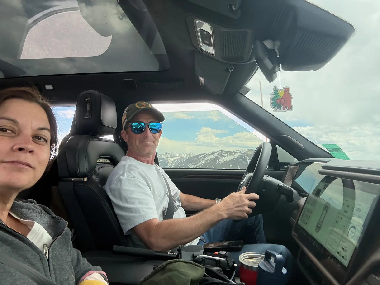 Selfie of a couple inside the Rivian R1T cab on Trail Ridge Road, a cartoon bear air freshener hanging from the mirror and snow-capped peaks through the windshield