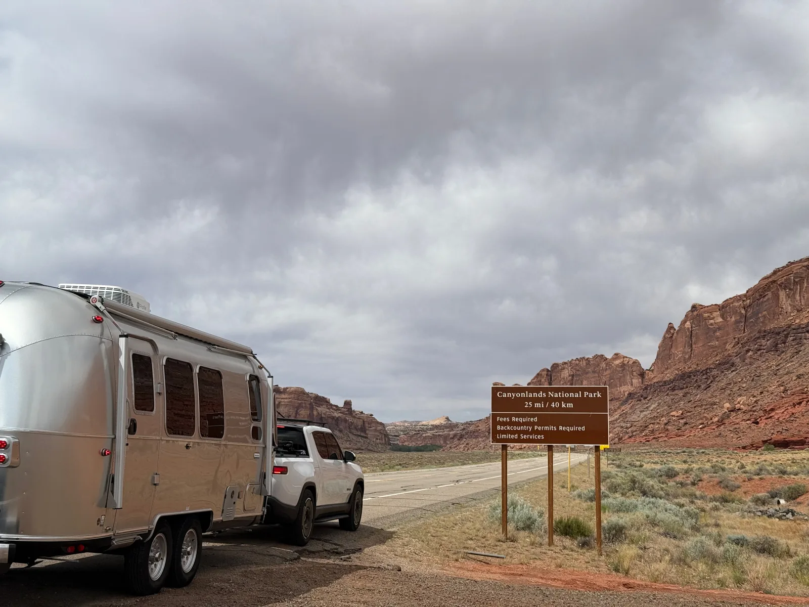 The Airstream and Rivian R1T parked beside the brown Canyonlands National Park entrance sign under moody Utah clouds
