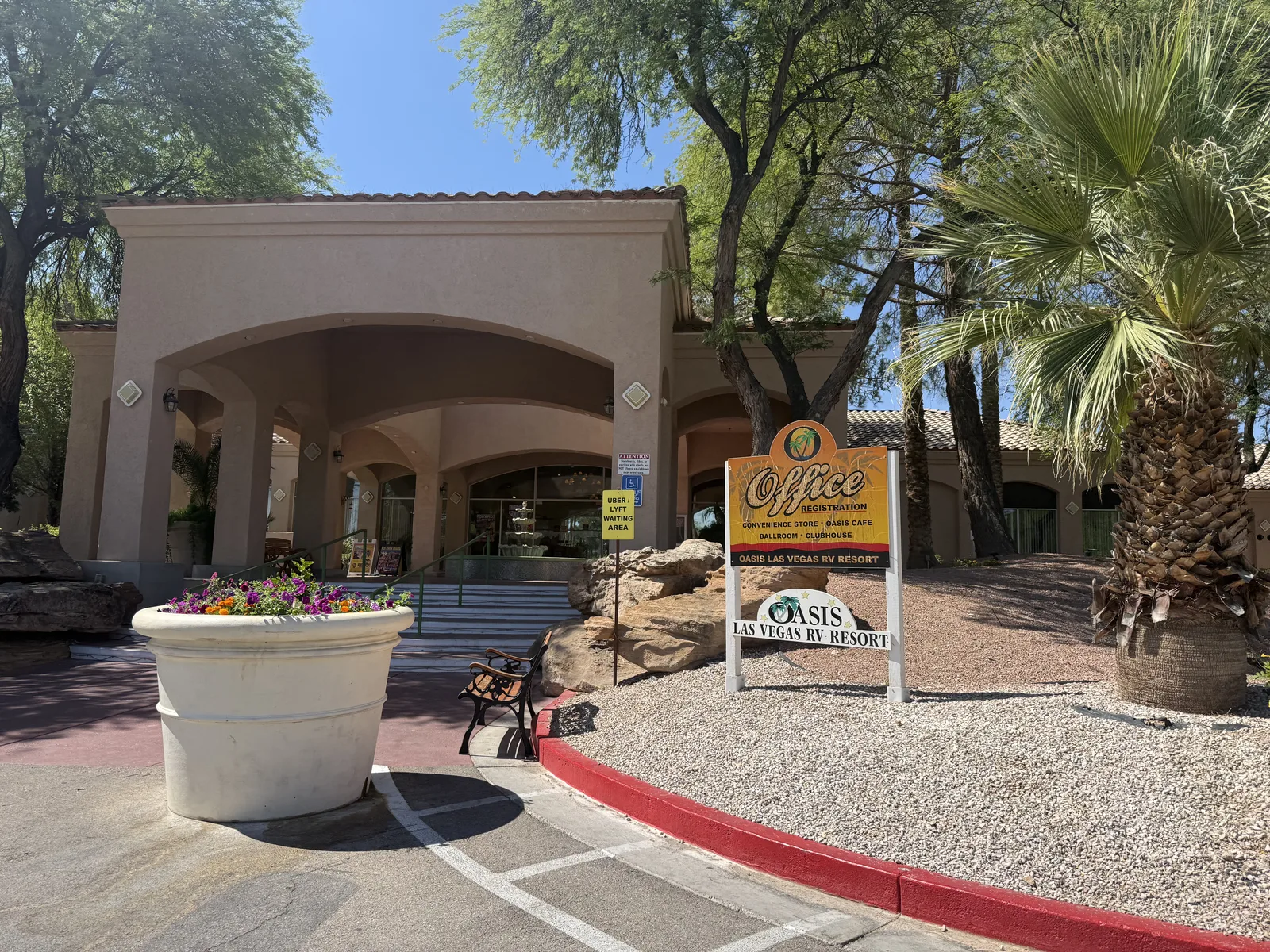 The arched stucco office and entrance sign of the Oasis Las Vegas RV Resort under tall palms and a clear blue desert sky on the final morning