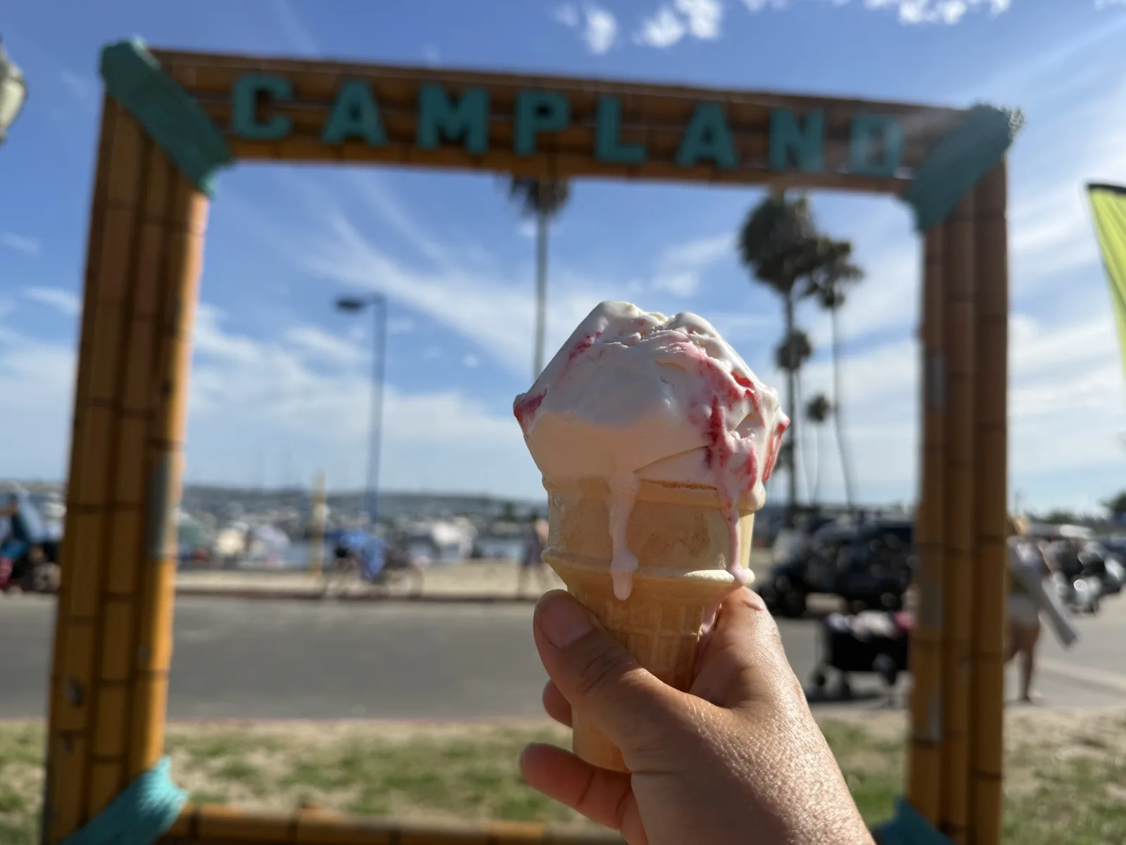A soft-serve cone held up inside the wooden CAMPLAND photo frame at Campland on the Bay, the Mission Bay marina and palms framed behind it