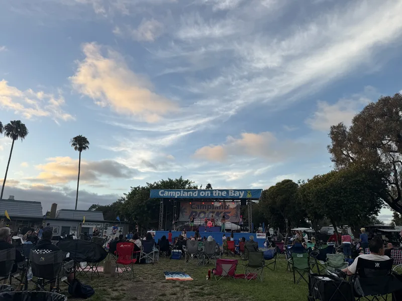 The concert lawn at Campland on the Bay at dusk, campers in folding chairs facing the Campland ROCKS stage with palms and a streaked sunset sky behind
