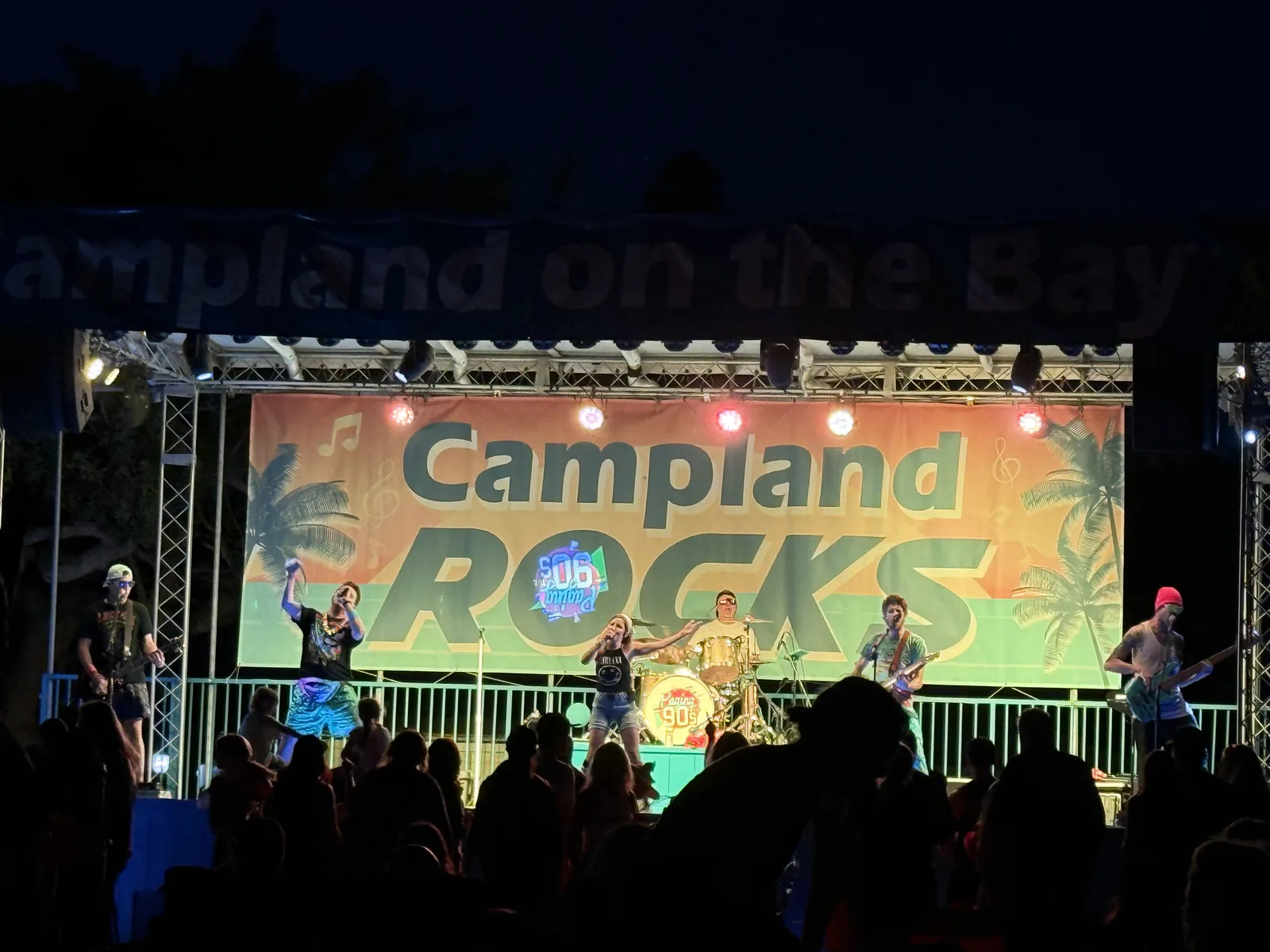 A band on the Campland ROCKS stage after dark at Campland on the Bay, stage lights on the players and the crowd in silhouette under the Campland on the Bay banner