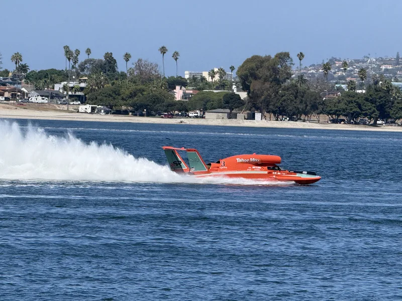 An orange unlimited hydroplane throwing a rooster tail across Mission Bay during the boat races, seen from the Fiesta Island shore with the Pacific Beach hills behind