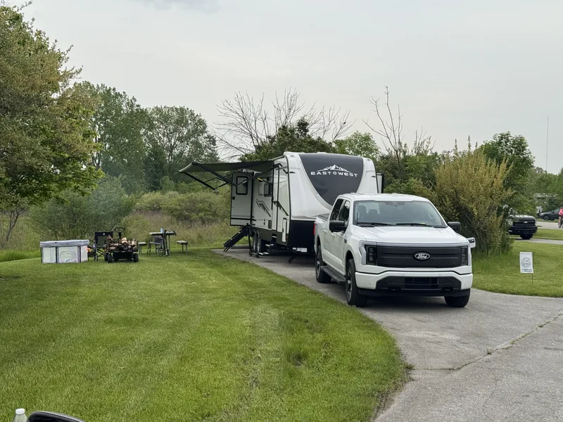 The East to West Alta with its awning extended and the white Ford F-150 Lightning parked alongside at a grassy campsite.
