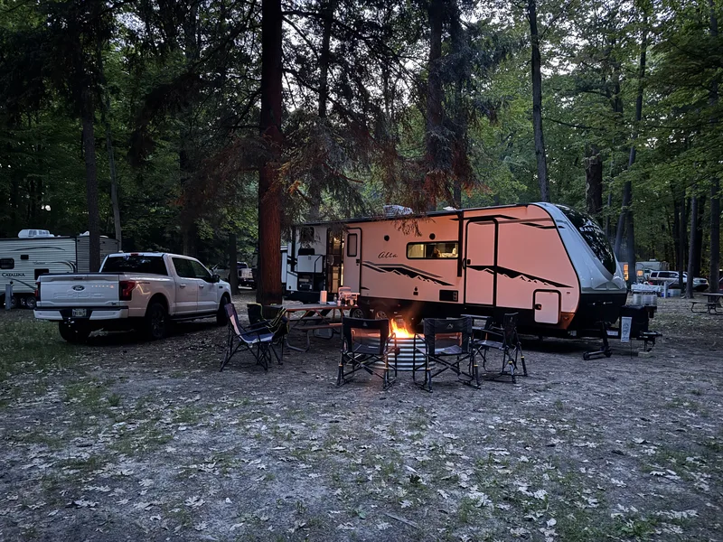 Dusk at a forested campsite, a campfire lit, the East to West Alta and Ford F-150 Lightning tucked in among the trees.