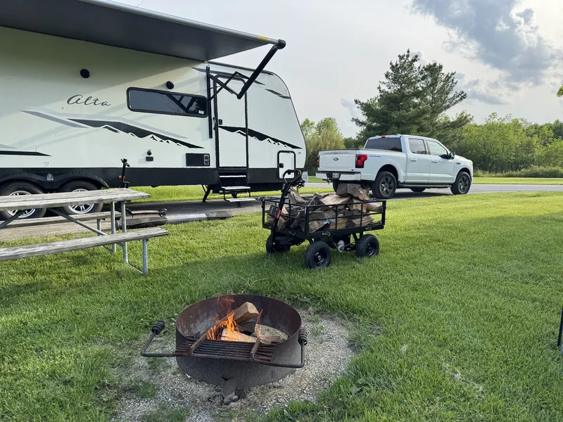 A firewood cart and a fresh fire in the ring at a grassy campsite, the East to West Alta and Ford F-150 Lightning behind.