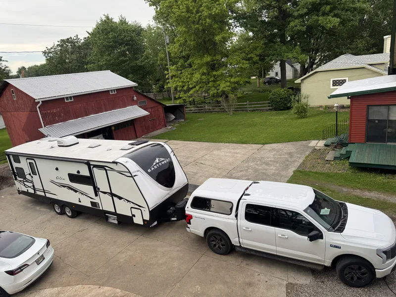 An elevated view of the Ford F-150 Lightning with a bed topper hitched to the East to West Alta in a driveway, a red barn behind.