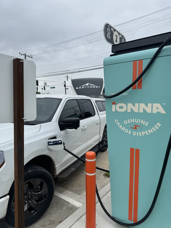 The Ford F-150 charge port connected at a teal IONNA fast charger, the East to West trailer in the background.