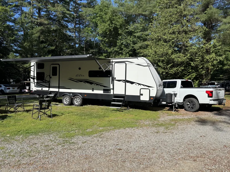 The East to West Alta with awning out and the white Ford F-150 Lightning parked among tall pines at a campsite.