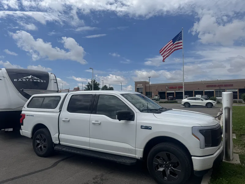 Side profile of the white Ford F-150 Lightning with a bed topper towing the East to West Alta, an American flag flying behind.